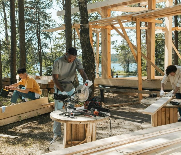 Team of diverse workers engaged in carpentry at a forested lakeside construction site.